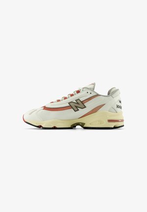Baskets New Balance avec une tige en cuir blanc, des accents rouges et beiges, une semelle extérieure en caoutchouc, une semelle intermédiaire rembourrée et un logo visible sur le côté.