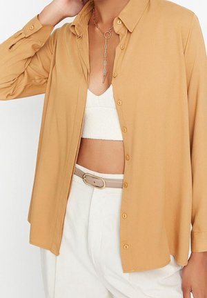 Vrouw draagt een lichtbruin overhemd met knopen over een witte crop top, witte broek met hoge taille en een beige riem, en een kettingketting.