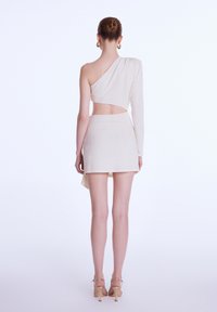 Robe asymétrique en tissu stretch beige clair avec une manche longue, détail découpé à la taille et une jupe courte. Texture lisse.