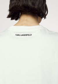 Bílý bavlněný tričko s kulatým výstřihem. Na zadní straně je umístěn text "KARL LAGERFELD" v černé barvě, vycentrovaný blízko límce.