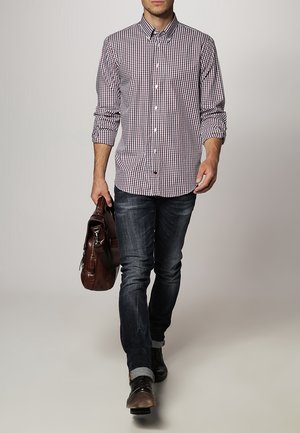 Mężczyzna idący, ubrany w czerwoną i białą koszulę w kratę, ciemne podwinięte dżinsy, czarne buty, trzymający w prawej ręce brązową skórzaną torbę.