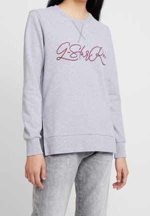 Kvinde iført lysegrå sweatshirt med lange ærmer og bordeaux kursivskrift samt matchende vaskede grå jeans, stående mod hvid baggrund.