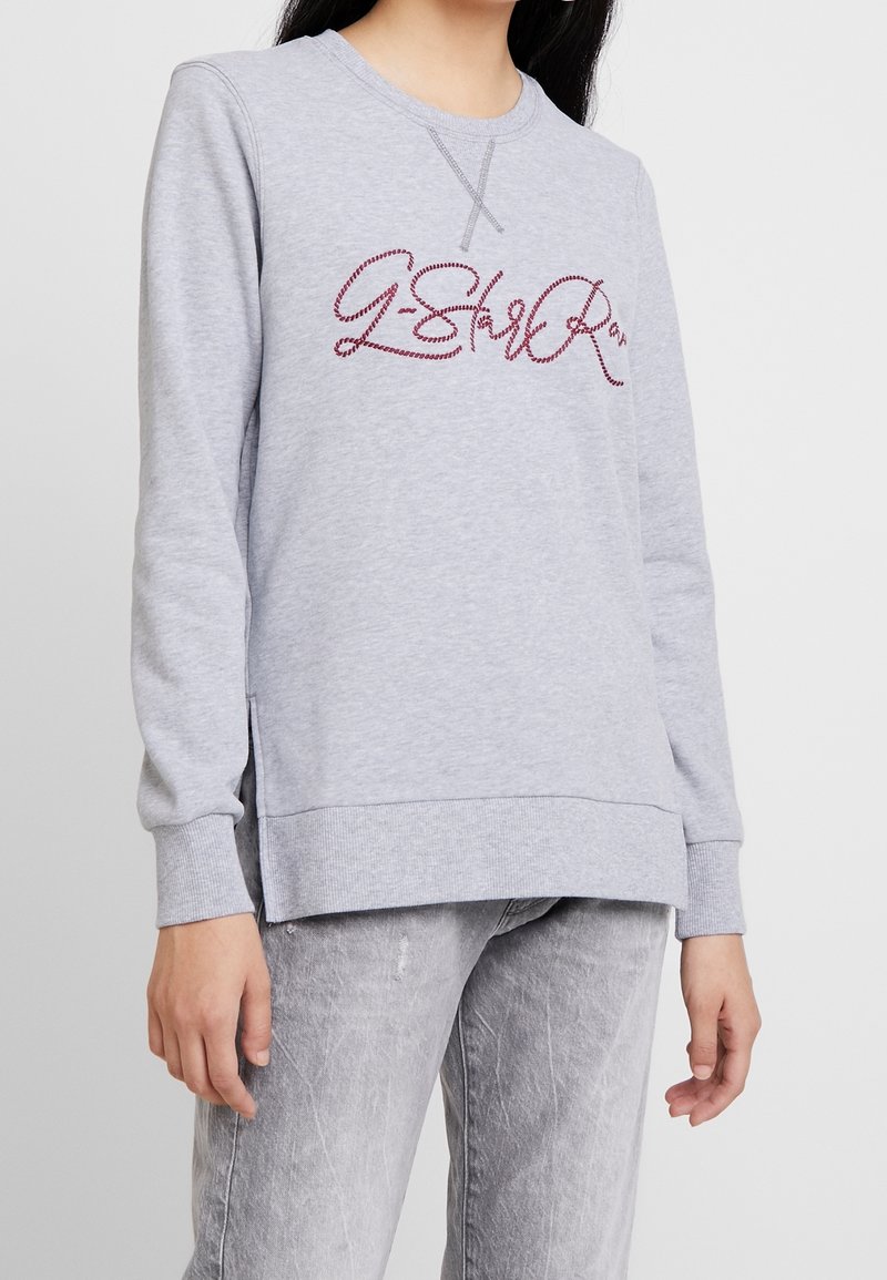 Femme portant un sweat-shirt gris clair à manches longues avec des lettres cursives bordeaux et un jean gris délavé assorti, debout sur un fond blanc.