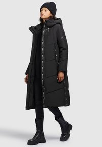 Langer schwarzer Puffer-Mantel mit Kapuze, ausgestattet mit einem Reißverschluss und Druckknopfverschluss, seitlichen Reißverschlusstaschen und einem gemusterten Innenfutter. Über schwarzer Kleidung getragen.