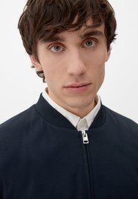 Granatowa kurtka bombowca o gładkiej teksturze, z zapięciem na zamek błyskawiczny i okrągłym kołnierzem, noszona na białej koszuli z kołnierzykiem. Prosty, nowoczesny design.