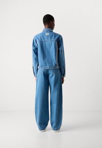 Giacca di jeans azzurro chiaro con cuciture visibili, chiusura a bottoni e logo sul retro. Pantaloni in denim a gamba larga con tasche sottili e vestibilità rilassata.