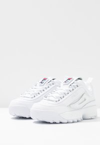 Scarpe da ginnastica bianche con un design robusto, tomaia in pelle e mesh testurizzato, accenti grigi e una suola in gomma spessa. Presenta il logo sulla linguetta.