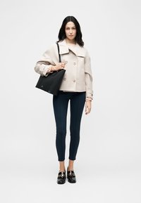 Naine, kes kannab beezi kahekordset jopet, meresiniseid kitsaid pükse, musti loafer-type jalatseid ning kannab suurt musta käekotti õlal.