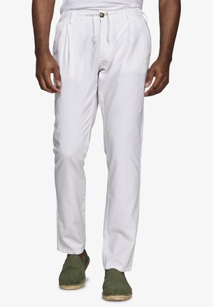 Mężczyzna w białych plisowanych spodniach z guzikiem i szlufkami na pasek, w ciemnozielonych butach wsuwanych, stoi na białym tle.