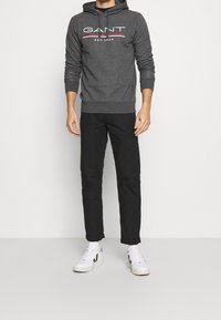 Uomo che indossa un hoodie GANT grigio scuro, pantaloni neri, calzini bianchi e sneakers bianche con dettagli neri, in piedi contro uno sfondo bianco uniforme.
