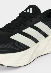 Chaussure de sport noire et blanche avec des lacets noirs, une semelle blanche et trois bandes blanches sur le côté, montrant les détails vus de côté et de dessus.