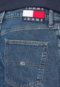 Närbild av blå denimjeans med bakficka och Tommy Jeans-märke ovanför fickan samt liten flagglogotyp på fickan.