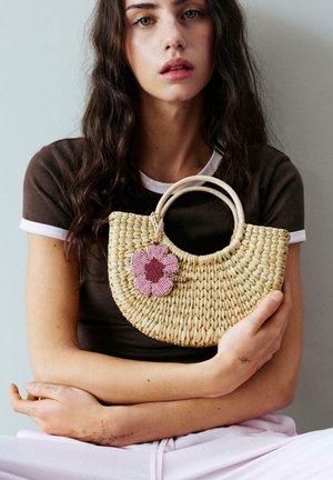 Jeune femme aux longs cheveux tient un sac à main en paille tressée décoré d'une fleur rose en perles, portant un t-shirt marron et un pantalon clair.