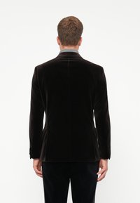 Homem de costas, vestindo um blazer de veludo escuro sobre uma camisa cinza, contra um fundo branco liso.