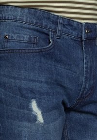 Vaqueros de mezclilla azul oscuro con un parche desgastado y distress, cierre de botón y bolsillos laterales. Tejido de algodón con una textura suave.