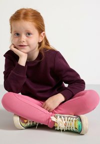 Burgunderrød sweatshirt, lyserøde leggings og iriserende højhælede sneakers. Håret er rødt og bølget, med fregner tydeligt synlige i ansigtet.