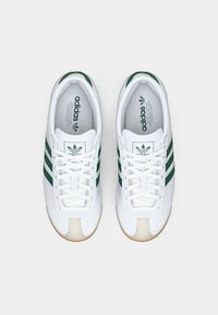 Zapatillas blancas con franjas verdes, parte superior de piel lisa, puntera redonda, diseño con cordones, cuello acolchado y suela de goma con acentos beiges.