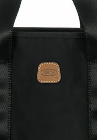 Zwarte stoffen draagtas met een gladde textuur, voorzien van een bruine leren patch met gestikte details waarin de merknaam "BRIC'S" is opgenomen.