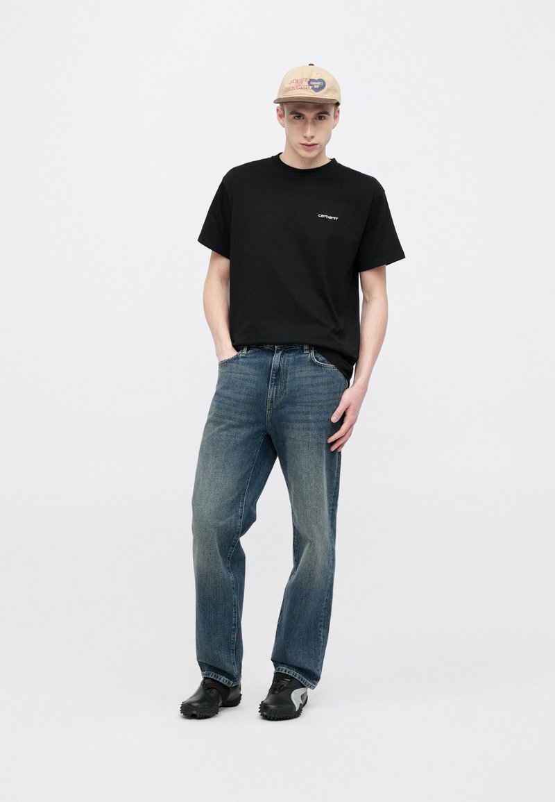 Black cotton t-shirt with logo on the chest, paired with blue straight-leg denim jeans and black slip-on shoes. Beige cap with graphic detail.