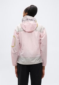 Άτομο με ροζ και γκρι μπουφάν Columbia με κουκούλα, γυρισμένο ανάποδα, εμφανίζοντας το λογότυπο "Columbia" στο γιακά και τσέπη από δίχτυ στο αριστερό μανίκι.
