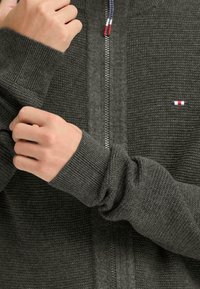 Grå zip-up jakke med varierte teksturer, med ribbestrikkede mansjetter og en liten logo i rød, hvit og blå på venstre side.