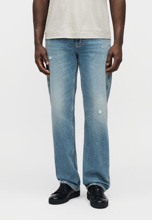 Jean bleu clair à jambes droites avec une texture délavée et des détails usés. Associé à des baskets noires. Design minimaliste, style cinq poches.