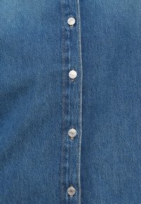 Μπλε πουκάμισο από τζιν με κουμπιά στο μπροστινό μέρος που διαθέτουν ασημί κουμπιά, αντίθετο ράψιμο και ελαφριά υφή στο ύφασμα.