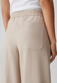 Pantalons beige, coupe décontractée avec une jambe large et une taille élastique. Présente une poche arrière unique avec un rabat rectangulaire. Texture douce.