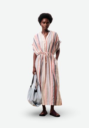Vrouw staand, gekleed in een lange jurk met verticale meerkleurige strepen en een strikceintuur, ze draagt een grote stoffen tas en donkere instapsandalen.