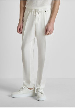 Persona con pantalones blancos con cordón y zapatillas blancas, de pie contra un fondo claro y sencillo.