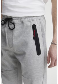 Light gray joggers with a textured fabric, featuring a black zippered pocket accented with red pull tab and an elastic waistband with drawstring.