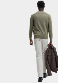 Maglione verde oliva, design aderente, scollatura a girocollo, maglia testurizzata. Pantaloni chiari, vestibilità rilassata. Tiene in mano una giacca marrone scuro. Scarpe nere.