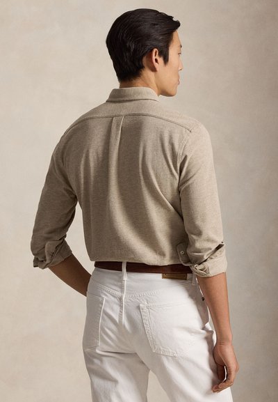 Moški, ki nosi bež majico z dolgimi rokavi, zavihanimi rokavi, zataknjeno v bele hlače z rjavim pasom, prikazan od zadaj.