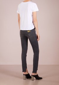 Mujer vestida con una camiseta blanca de manga corta, jeans ajustados oscuros grises con una etiqueta dorada en el bolsillo trasero y zuecos negros con tacones de bloque de madera.