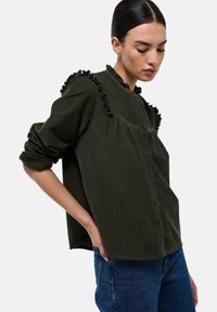 Chemise en velours côtelé vert olive avec des accents froncés le long du col et des épaules ; manches longues avec des poignets retroussés.