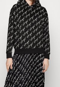 Sudadera negra con capucha y falda plisada con un patrón de texto blanco en diagonal escrito a mano que dice "Hugo", lucida por una persona con las manos a los lados.