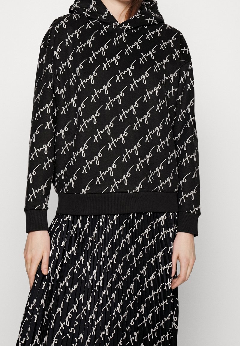 Sudadera negra con capucha y falda plisada con un patrón de texto blanco en diagonal escrito a mano que dice "Hugo", lucida por una persona con las manos a los lados.