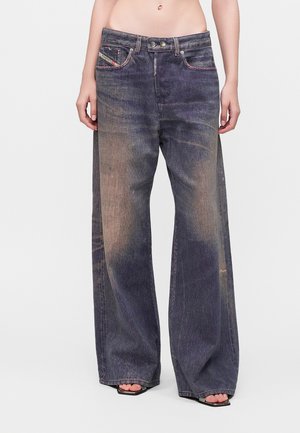 Παντελόνι τζιν από μαύρο ξεθωριασμένο denim με φαρδιά μπάτζα, φορεμένο με μαύρα σανδάλια ανοιχτών δακτύλων, απεικονίζεται σε ένα άτομο από τη μέση ως τα πόδια σε απλό φόντο.