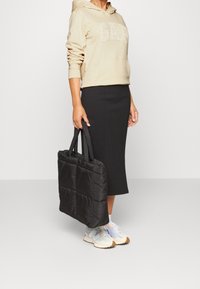 Zwarte gewatteerde totebag met dubbele handvatten, voorzien van een doorgestikt ontwerp. Gecombineerd met een beige hoodie en een zwarte rok tot aan de knieën.