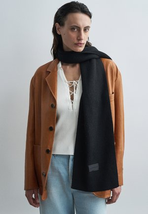 Černý žebrovaný šátek přehozený přes světle hnědou koženou bundu, pod kterou je bílý strukturální top, se subtílní štítkovou nálepkou na okraji šátku.