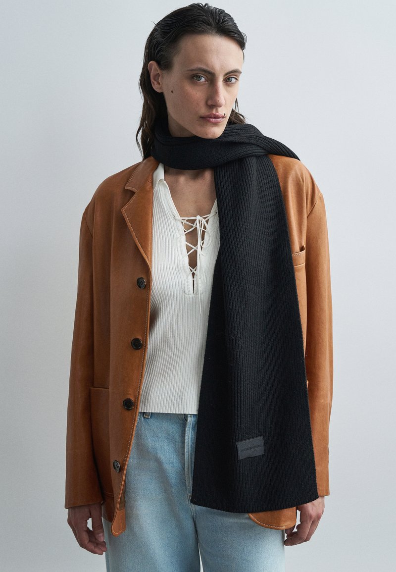Black ribbed scarf draped around a light brown leather jacket over a white textured top, with a subtle logo tag on the scarf's edge.