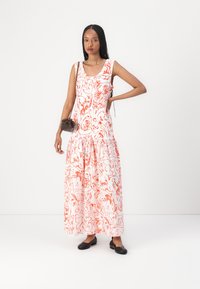 Langes, weißes Kleid mit roten abstrakten Mustern, ärmelloses Design und geraffter Taille. Kombiniert mit einer kleinen braunen Handtasche und schwarzen Ballerinas.