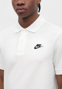 Hombre vestido con una camiseta polo blanca de Nike con un logo negro en el pecho izquierdo, visible desde los hombros hasta la mitad del torso sobre un fondo liso.