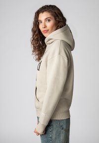 Beige hoodie met een voorkraag, geribbelde manchetten en zoom, met zwarte tekst op de voorkant en een logodetail op de mouwen.