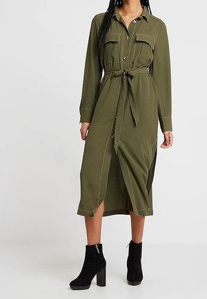 Vrouw die een olijfgroen midiklän met knopen draagt, met borstzakken, een taillekoord en zwarte enkellaarsjes met hak, staand tegen een effen achtergrond.