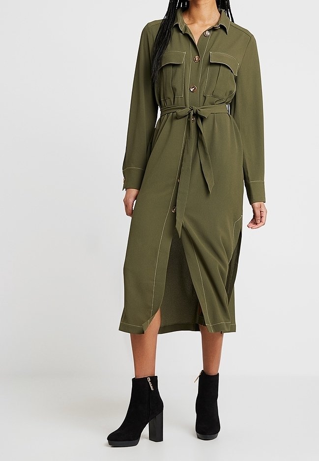 Femme portant une robe midi boutonnée vert olive avec poches poitrine, ceinture à nouer, et bottines à talons noires, debout sur un fond uni.