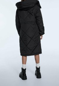 Czarna pikowana kurtka puchowa z kapturem, zwężona w talii i z długimi rękawami. Ma długość do kolan i teksturowany materiał.