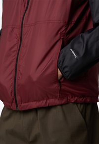 Giacca a vento rossa e nera, con zip sul davanti, tasche laterali, texture morbida, presenta il logo "WINDWALL" sulla manica. Abbinata a pantaloni marroni.