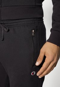 Sweatshirt noir avec des poignets côtelés et une poche zippée ; présente un petit logo brodé en rouge et blanc près de la poche. Texture lisse.