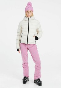 Witte pufferjack met capuchon, zwarte rits en elastische manchetten; roze broek; roze gebreide beanie met pompon; zwarte skibotten.
